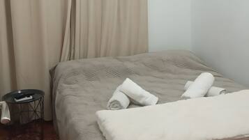 A bedroom with a bed, beige curtains, a small side table, and a patterned floor.