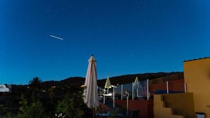 A night sky with stars, a plane in flight, outdoor seating with umbrellas, and a building with a modern design. - Vacation Home "Casa La Viña" with Sea View, Private Pool & Wi-Fi (Puntagorda)