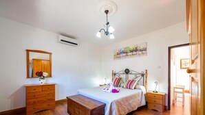 A bedroom with a wooden bed, a nightstand, a dresser, a bench, a painting on the wall, and an air conditioning unit. - Vacation Home "Casa La Viña" with Sea View, Private Pool & Wi-Fi (Puntagorda)