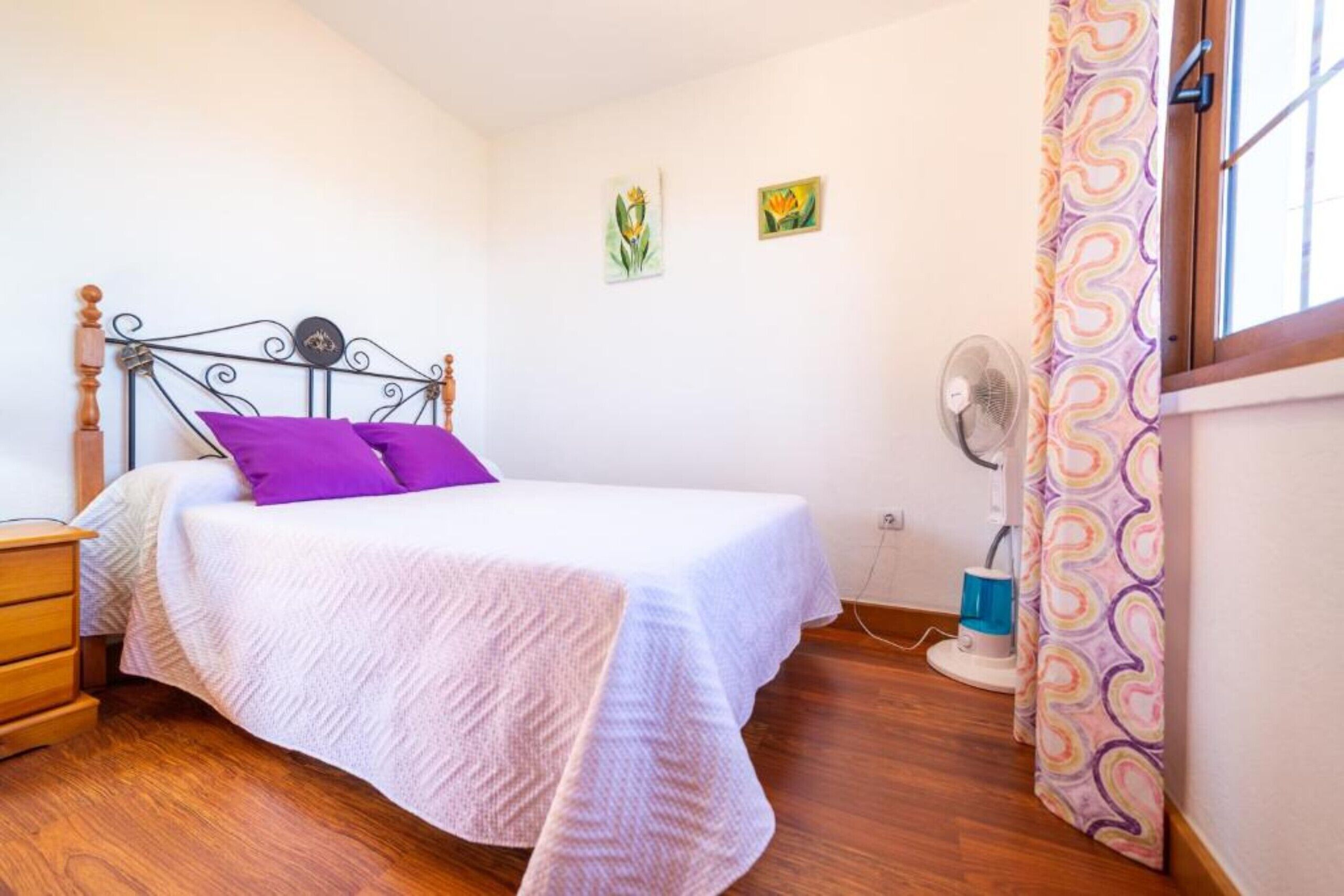 A bedroom with a bed, a nightstand, a wall-mounted fan, and a window with curtains.