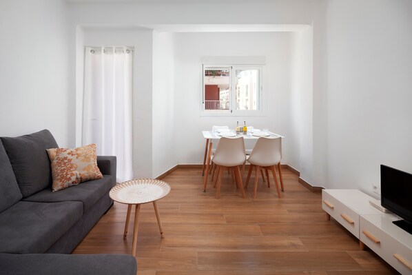 A living room with a sofa, a dining table with chairs, a TV, and a window with a view of buildings.