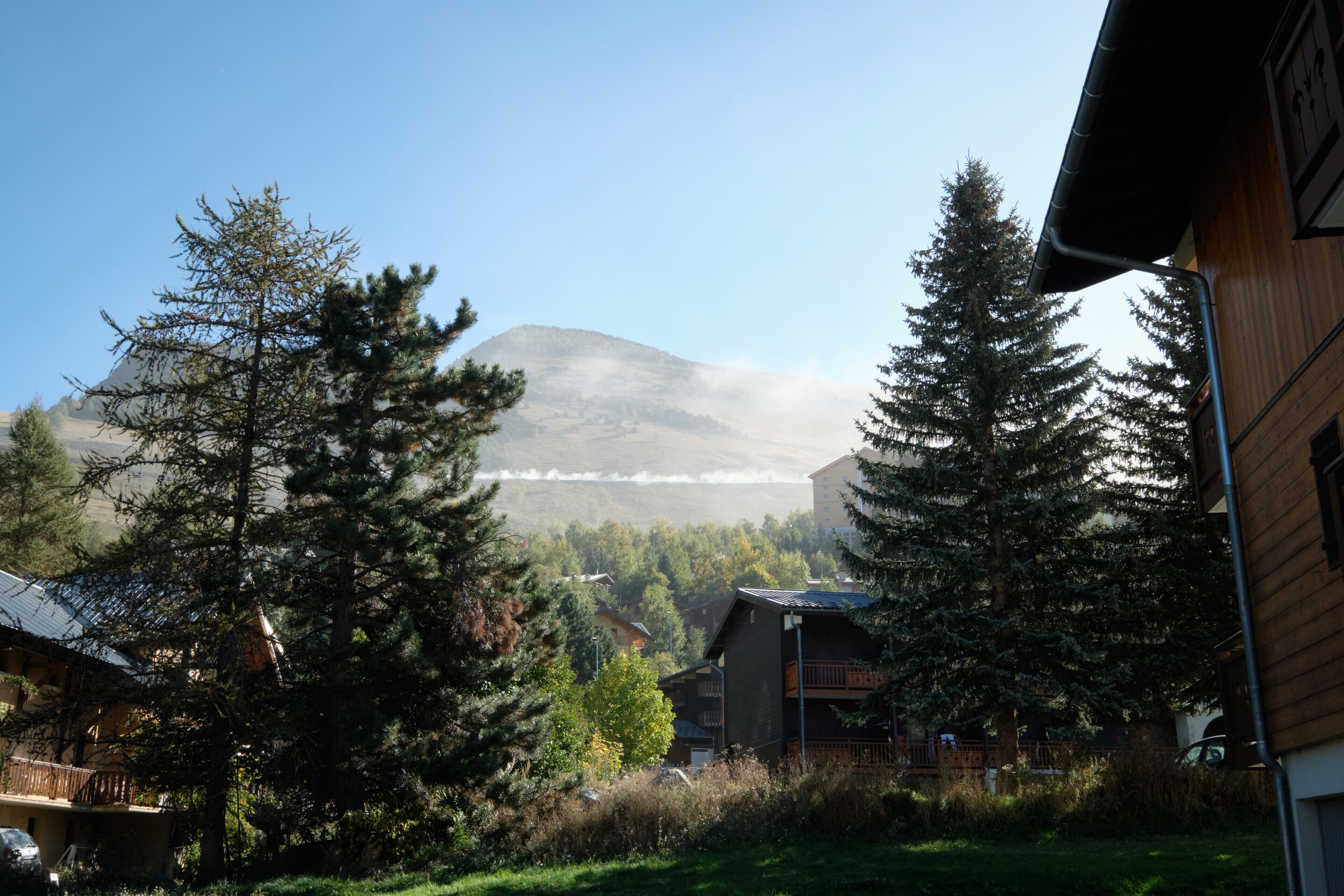 A mountain with a forested area in the foreground, a wooden building, and a clear sky.