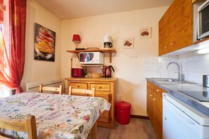 A compact kitchen with a microwave, kettle, and sink, wooden cabinets, a dining table with a floral tablecloth, and a red trash bin.