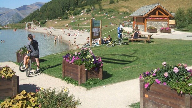 Un resort sul lago con una casetta di legno, una pista ciclabile e una zona verde con aiuole fiorite.
