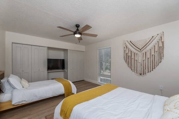 A bedroom with two beds, a ceiling fan, a TV, and a wall-mounted macrame decoration.
