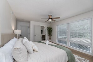 Un dormitorio con una cama grande, un ventilador de techo, una ventana con persianas y una puerta que da a otra habitación.