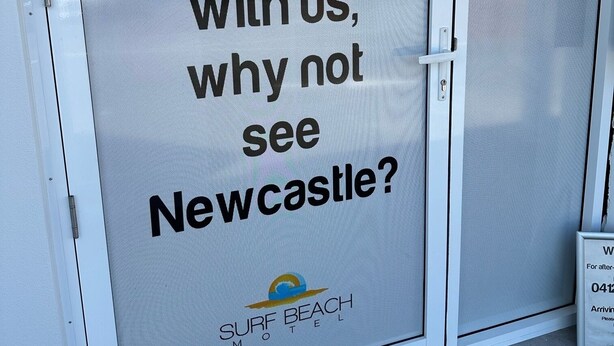 Una puerta con un cartel que dice, "Cuando se hospede con nosotros,  ¿por qué no conocer Newcastle?" y el logotipo del Surf Beach Motel.