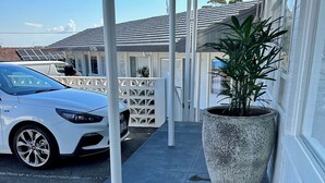 Un espacio exterior cubierto con un coche blanco estacionado cerca, una planta grande en maceta y una valla blanca.