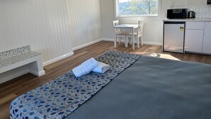 Una habitación moderna y bien iluminada, con un ventanal grande, una kitchenette pequeña, un comedor y una zona de estar con un banco y una alfombra estampada.