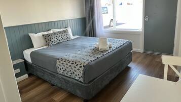 A neatly made bed with a patterned bedspread and pillows, a grey upholstered headboard, a window with curtains, and a door to the right.