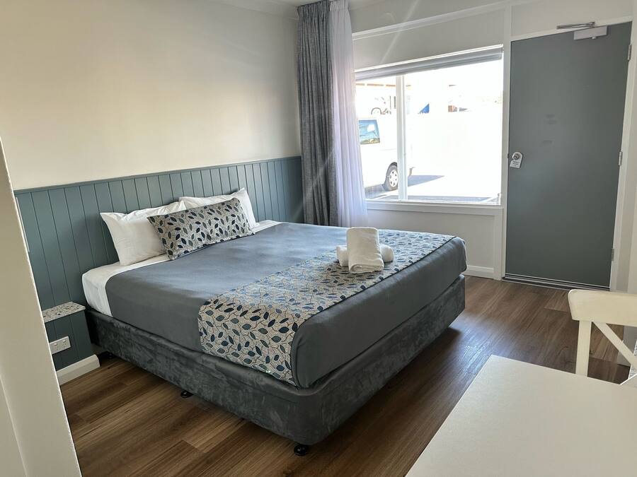 A neatly made bed with a patterned bedspread and pillows, a grey upholstered headboard, a window with curtains, and a door to the right.