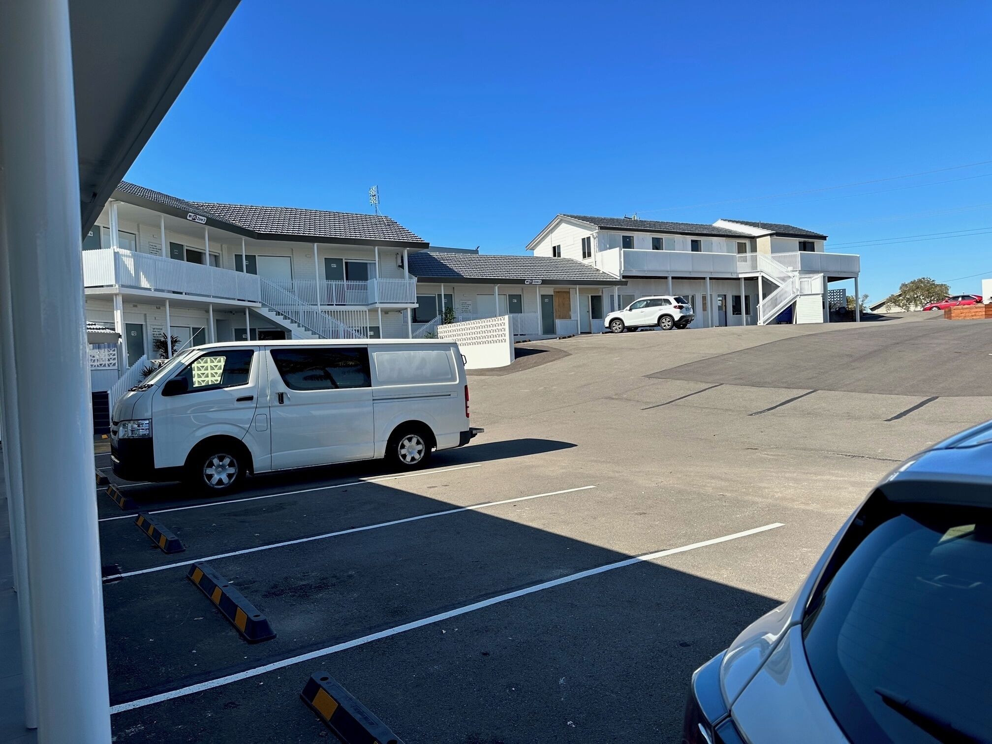 A parking lot with a white van and a white car, buildings with balconies and a staircase, and a clear blue sky.