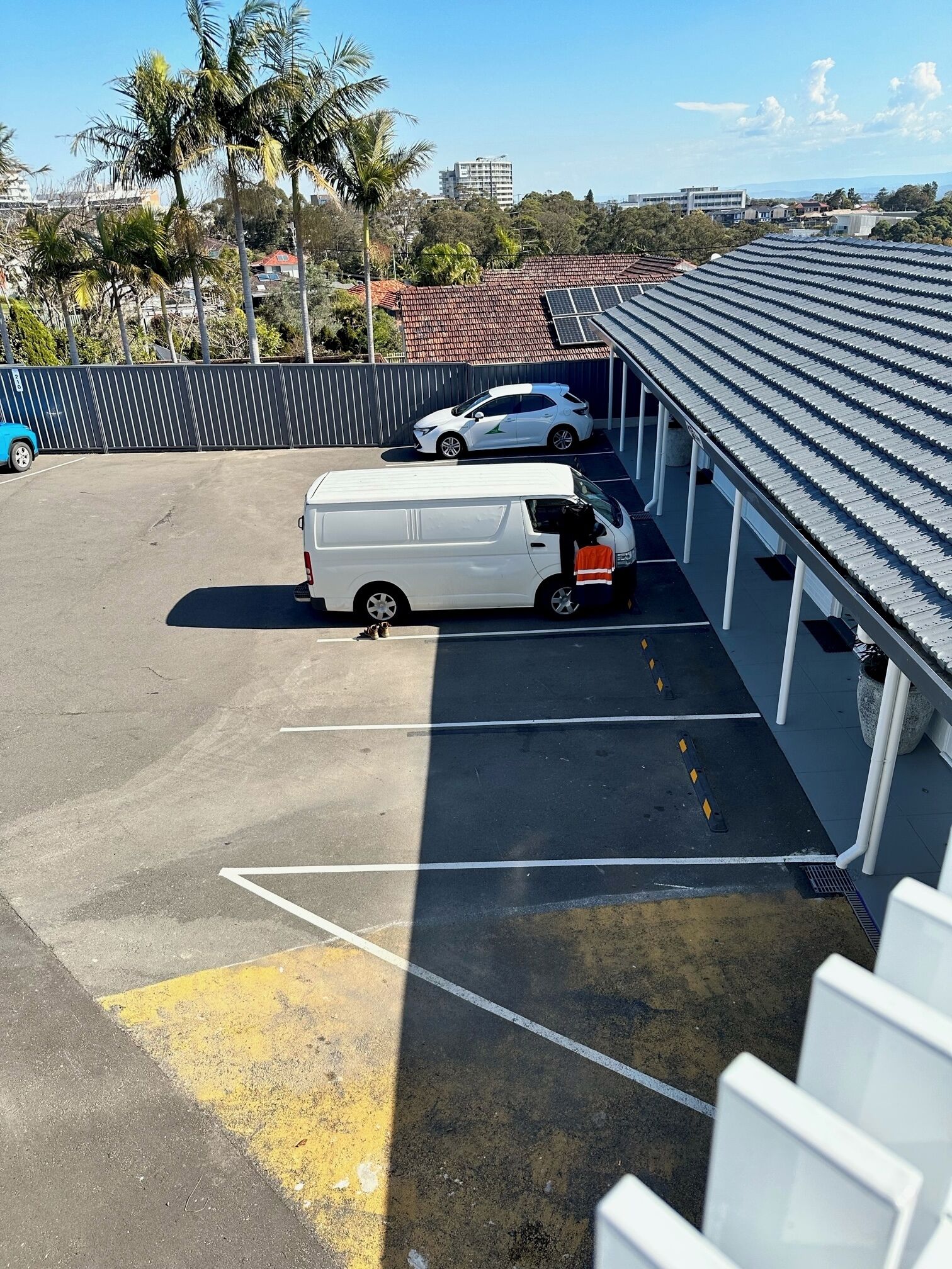 A parking area with a white van and a white car parked, a covered area with a tiled roof, and a view of buildings and trees in the background.