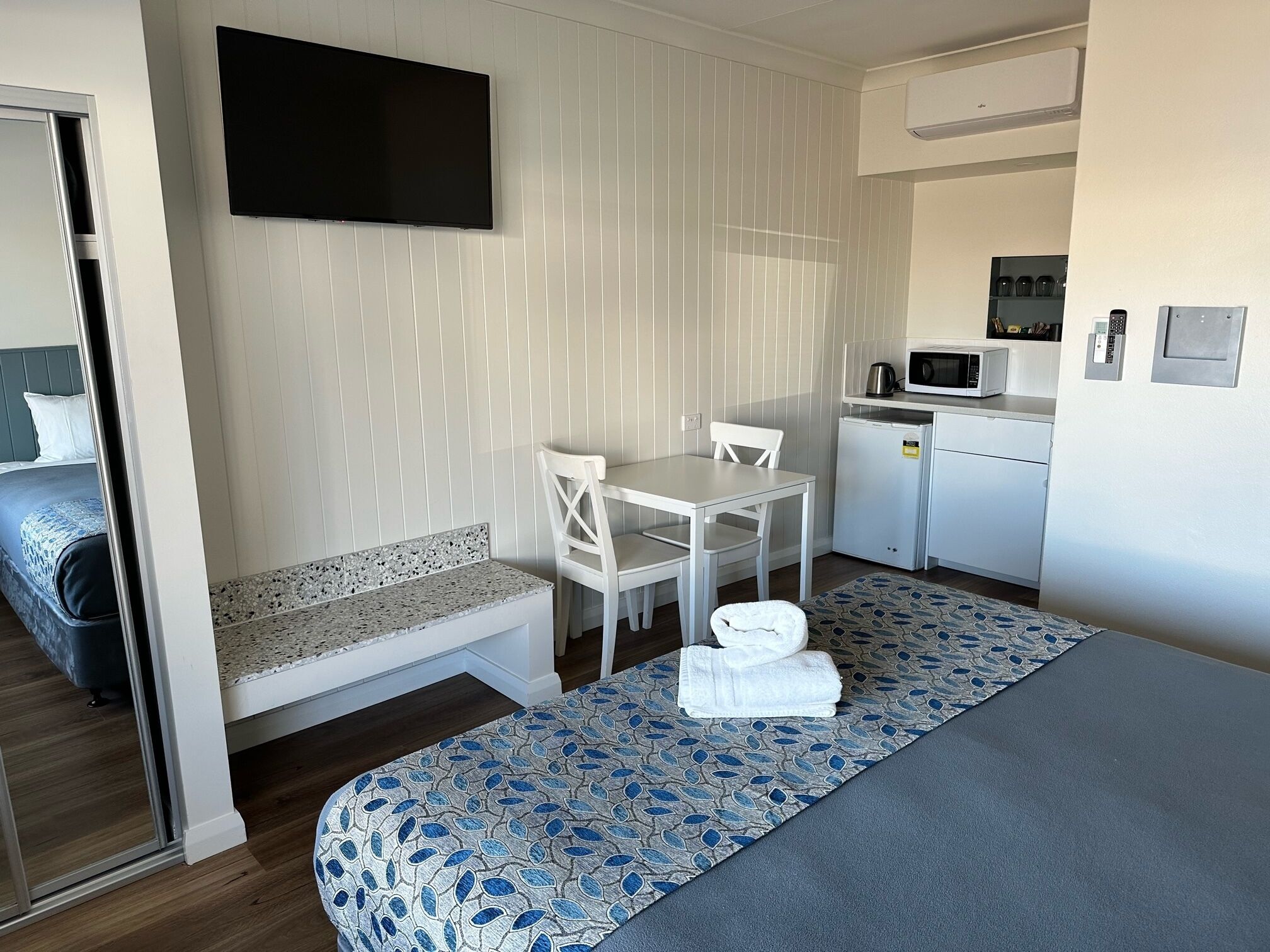 Habitació d'hotel senzilla amb llit, petita cuineta, zona de dinar i televisió penjada a la paret.