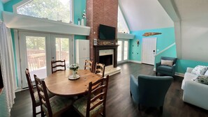 A spacious living area with a dining table, chairs, a fireplace, a TV mounted on the wall, and a staircase leading to an upper level.