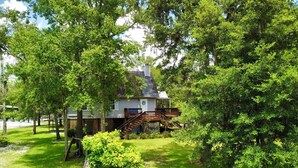 A house with a wooden deck and stairs leading to a porch, surrounded by lush greenery and trees.