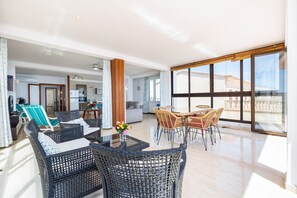 A spacious living area with wicker furniture, a dining table, and large windows offering a view of the outside.