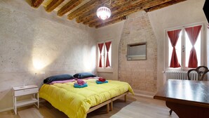 A bedroom with a bed, bedside table, wooden beams, brick wall, window with curtains, radiator, and a wooden bench. - Vacation Home "Ca' Lirica" with Wi-Fi & A/C (Venezia)