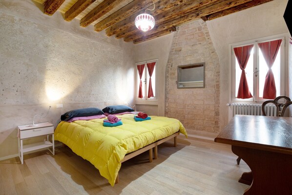 A bedroom with a bed, bedside table, wooden beams, brick wall, window with curtains, radiator, and a wooden bench.
