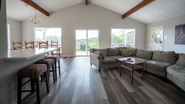 A spacious living area with a high ceiling, wooden beams, a large sofa, a dining area with chairs, and a kitchen counter. - Four bedroom with hot tub and views! (Kerrville)