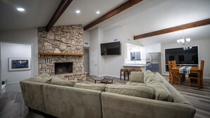 A living room with a stone fireplace, a sofa, a dining area with a table and chairs, and a kitchen in the background. - Four bedroom with hot tub and views! (Kerrville)