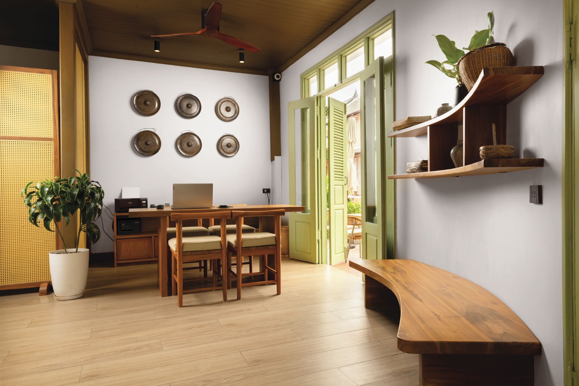 Une pièce comprenant une table à manger en bois, des chaises, un ordinateur portable, une plante en pot et un banc. On y trouve également des étagères murales ornées d’objets décoratifs et une fenêtre avec des volets verts.