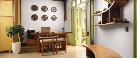 Una habitación con una mesa de comedor de madera, sillas, una computadora portátil, una planta en maceta y un banco. Hay repisas en la pared con adornos y una ventana con contraventanas verdes.