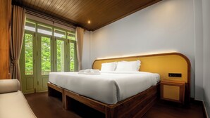 A bedroom with a wooden bed, a bench, a bedside table, and a view of greenery through the window.