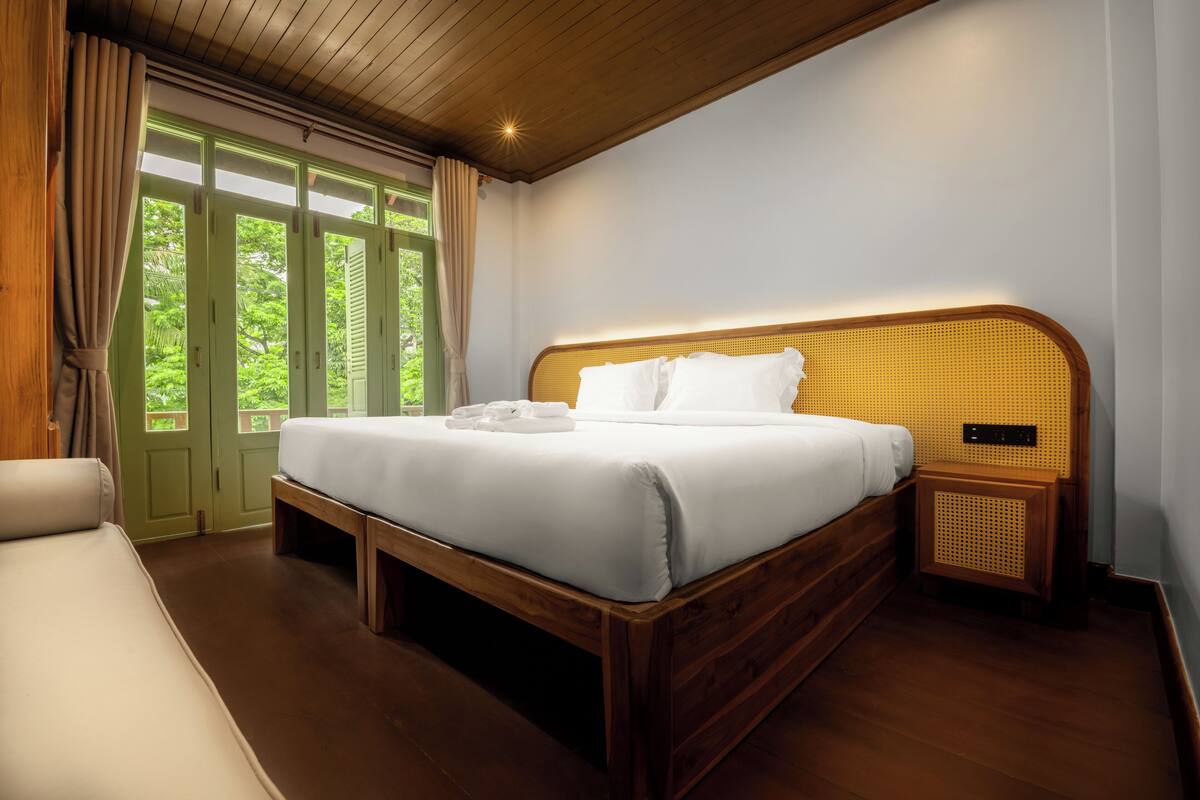 A bedroom with a wooden bed, a bench, a bedside table, and a view of greenery through the window.