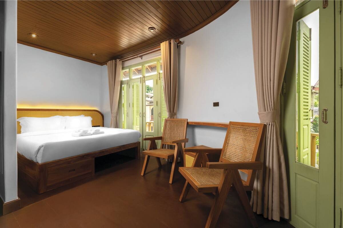 A hotel room with a wooden ceiling, a bed with white linens, two wicker chairs, and a view through a window with green shutters.