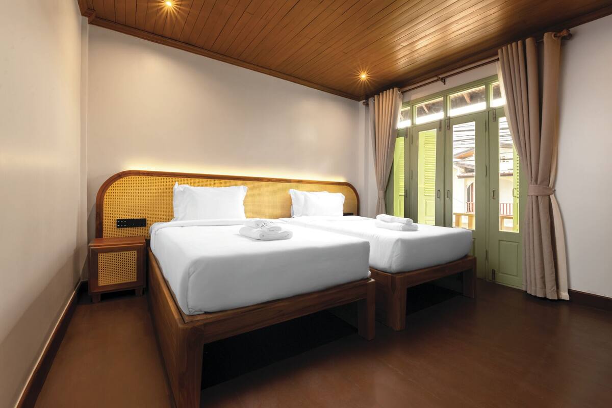 A hotel room with two wooden beds, a woven headboard, and a view of a green building through open shutters.
