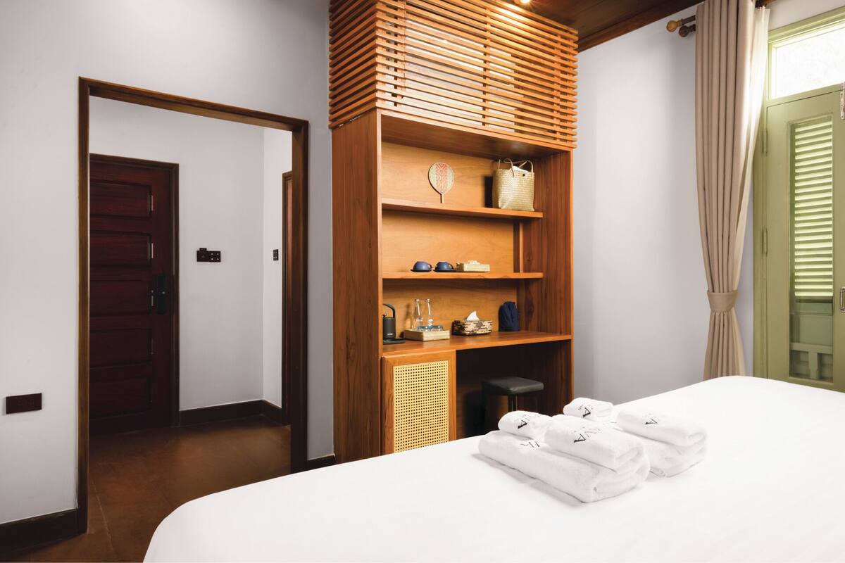 A hotel room with a bed, a wooden shelf with toiletries, a door, and a window with curtains.