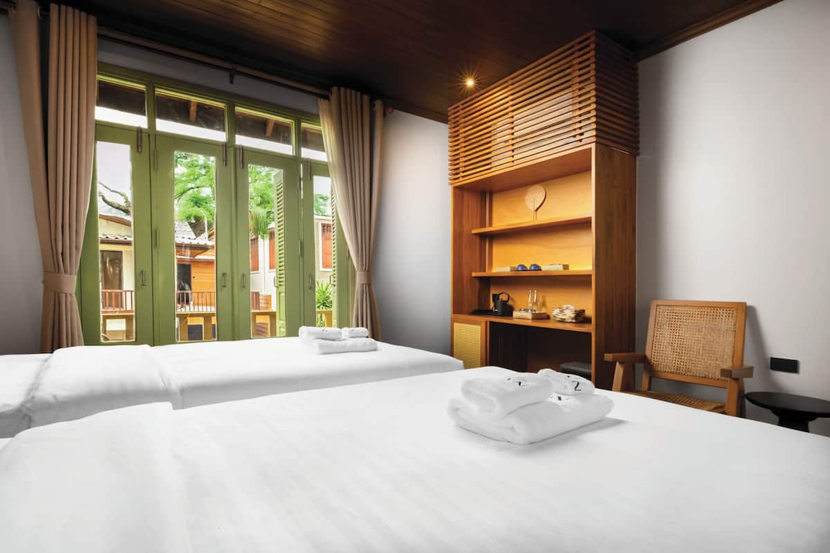 A hotel room with two beds, a wooden bookshelf, a chair, and a view of the outside.