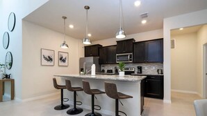 Een moderne keuken met donkere kastjes, een centraal eiland met barkrukken, hangende verlichting en een magnetron.