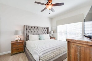 A bedroom with a tufted headboard, a bed with striped bedding, a wooden nightstand with a lamp, a wooden dresser, and a ceiling fan. - Private Pool, Prime Location, Beautiful Home (Kissimmee)