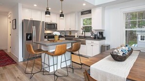 A modern kitchen with white cabinets, stainless steel appliances, a central island with bar stools, and a dining table with a white tablecloth. - Downtown Retreat with Bikes, Fire Pit & Kayaks (Saint Michaels)