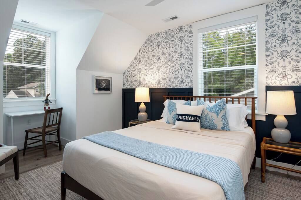 Une chambre à coucher avec un grand lit, deux tables de chevet avec des lampes, une chaise, un bureau et une fenêtre avec des stores.