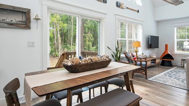 A spacious dining area with a wooden table and chairs, a large window with a view of trees, and a fireplace with a modern design. - Downtown Retreat with Bikes, Fire Pit & Kayaks (Saint Michaels)