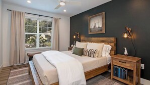 A bedroom with a large bed, a ceiling fan, a window with curtains, a nightstand with a lamp, and a framed picture on the wall. - Downtown Retreat with Bikes, Fire Pit & Kayaks (Saint Michaels)