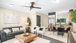Ein modernes Wohnzimmer mit einer Couch, Sesseln, einem Couchtisch und einem Essbereich mit Tisch und Stühlen. Es gibt einen Deckenventilator und Einbaulampen.