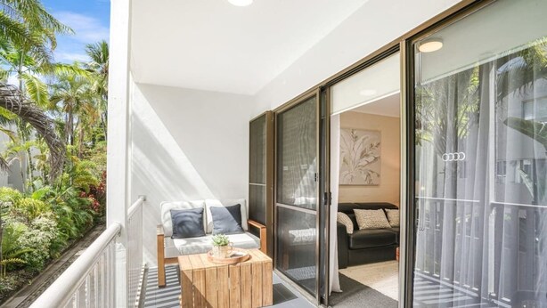 Ein Balkon mit einem Holztisch, ein weißes Sofa mit dunklen Kissen und ein Sofa im Inneren eines Raumes mit einem großen Fenster und einem gerahmten Kunstwerk.