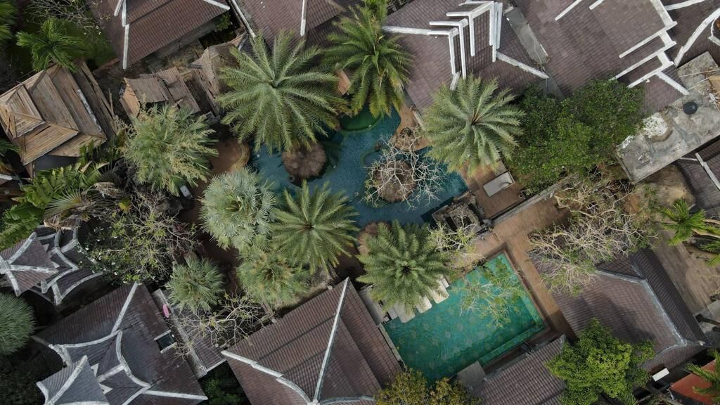 An aerial view of a courtyard with palm trees, a swimming pool, and a small bridge.