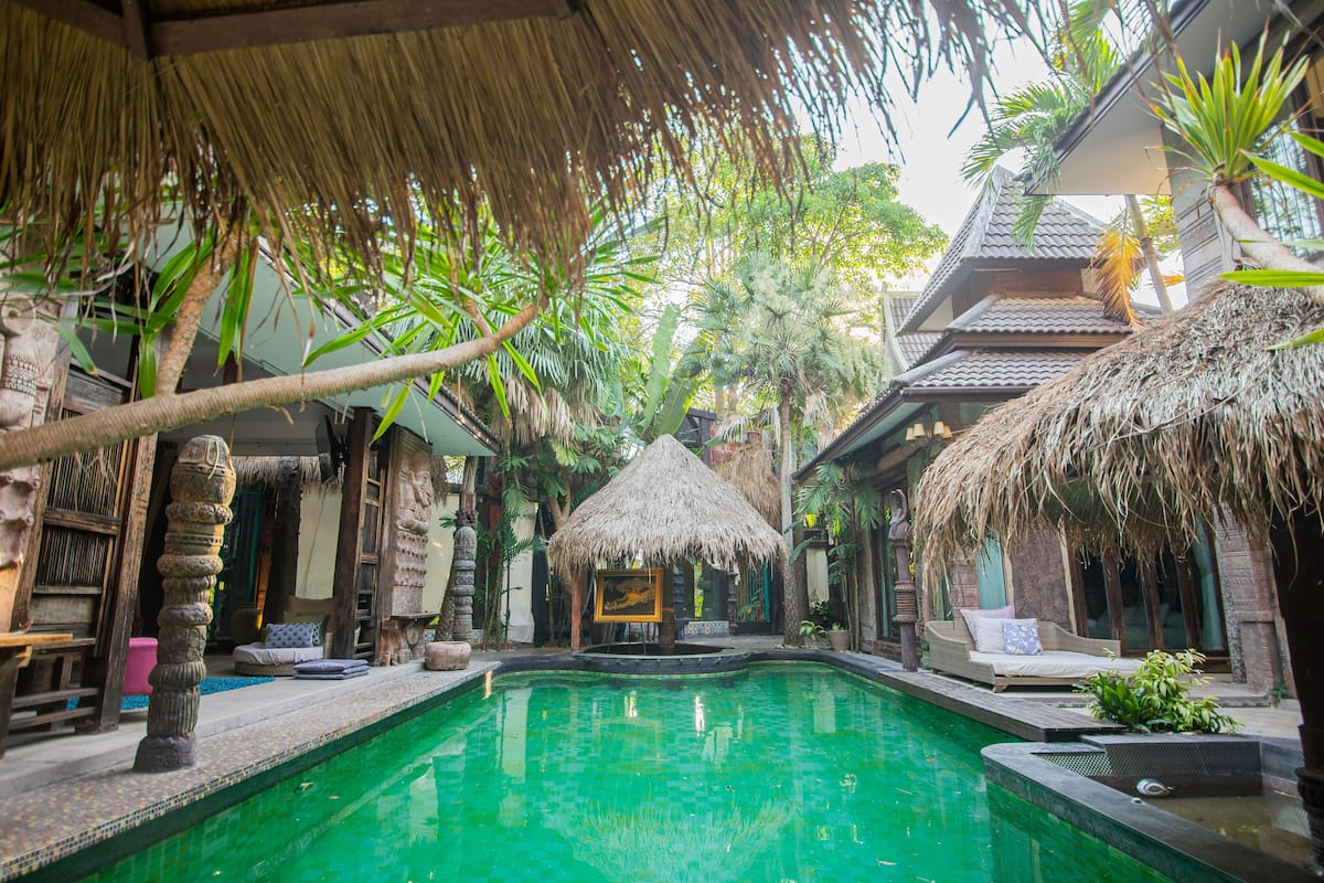 Un espace piscine agrémenté d’un toit de chaume, de mobilier en bois et de plantes tropicales.