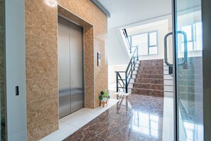 Un ascensor moderno con pared de mármol, una escalera con barandillas de metal y una planta en maceta en el suelo.