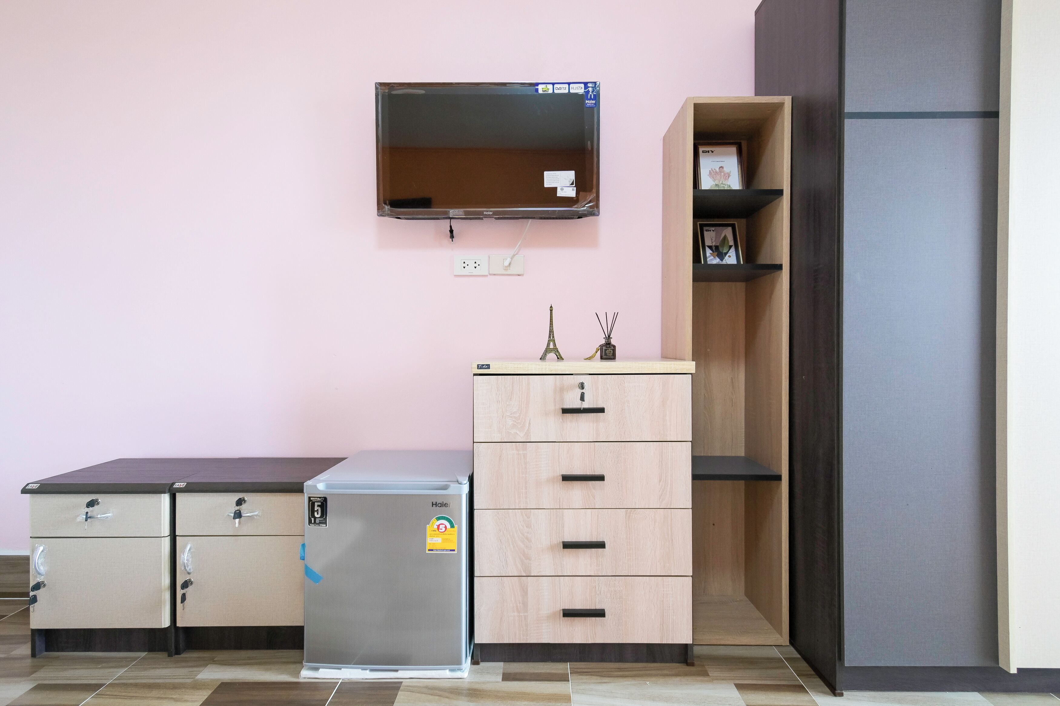 A compact kitchenette with a refrigerator, a small cabinet, a wooden shelf, and a wall-mounted TV.