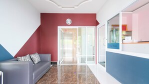 A modern interior with a grey sofa, a red accent wall, and a glass door leading to another room. - The Wish Hotel (Chachoengsao)