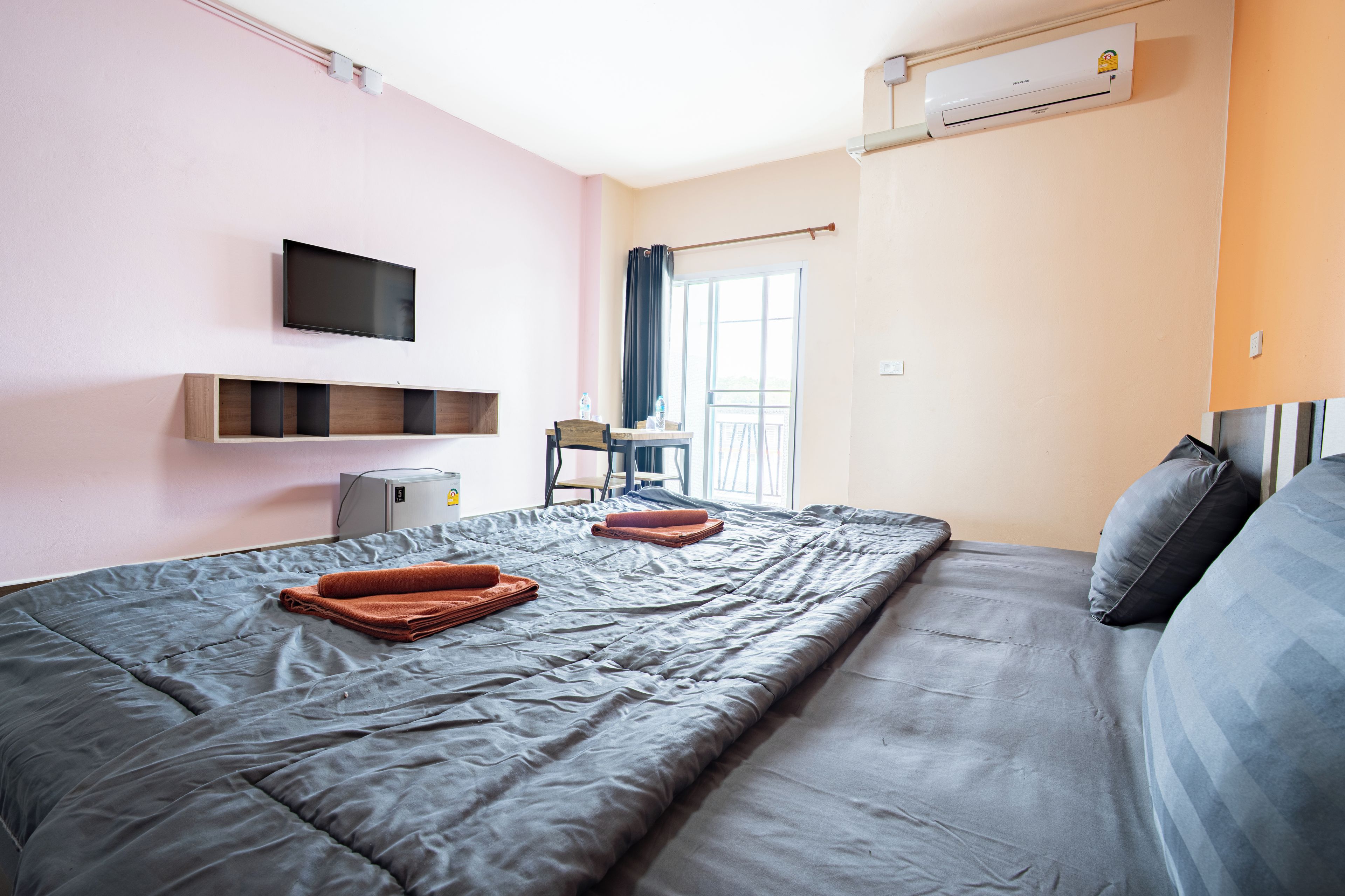 A hotel room with a bed, a flat-screen TV mounted on the wall, a small desk with a chair, and an air conditioning unit.