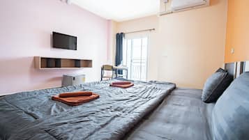 A hotel room with a bed, a flat-screen TV mounted on the wall, a small desk with a chair, and an air conditioning unit.