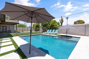 A rectangular swimming pool with a clear blue water surface, surrounded by a paved deck. There are lounge chairs and a large umbrella providing shade.