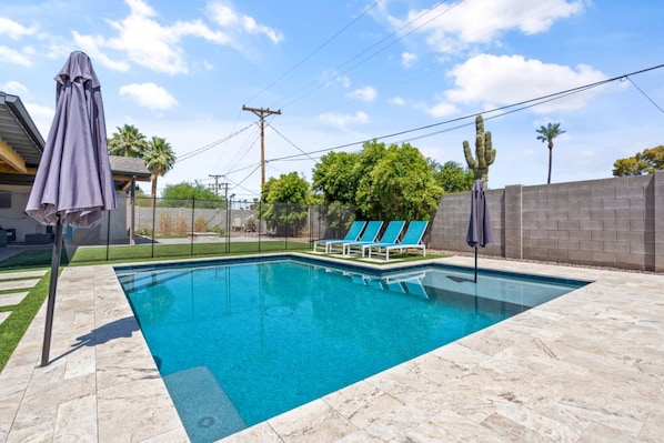 A rectangular swimming pool with a tiled deck, surrounded by a low wall and a fence. There are lounge chairs and umbrellas along the poolside.